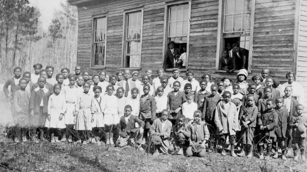 Black and white image of a large group of school children outside of their school building. Ages of students are varied.