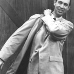 A grayscale half-body photograph of Fred Rogers putting his coat on to bid his audience goodbye at the end of an episode of the television show Mister Rogers' Neighborhood.