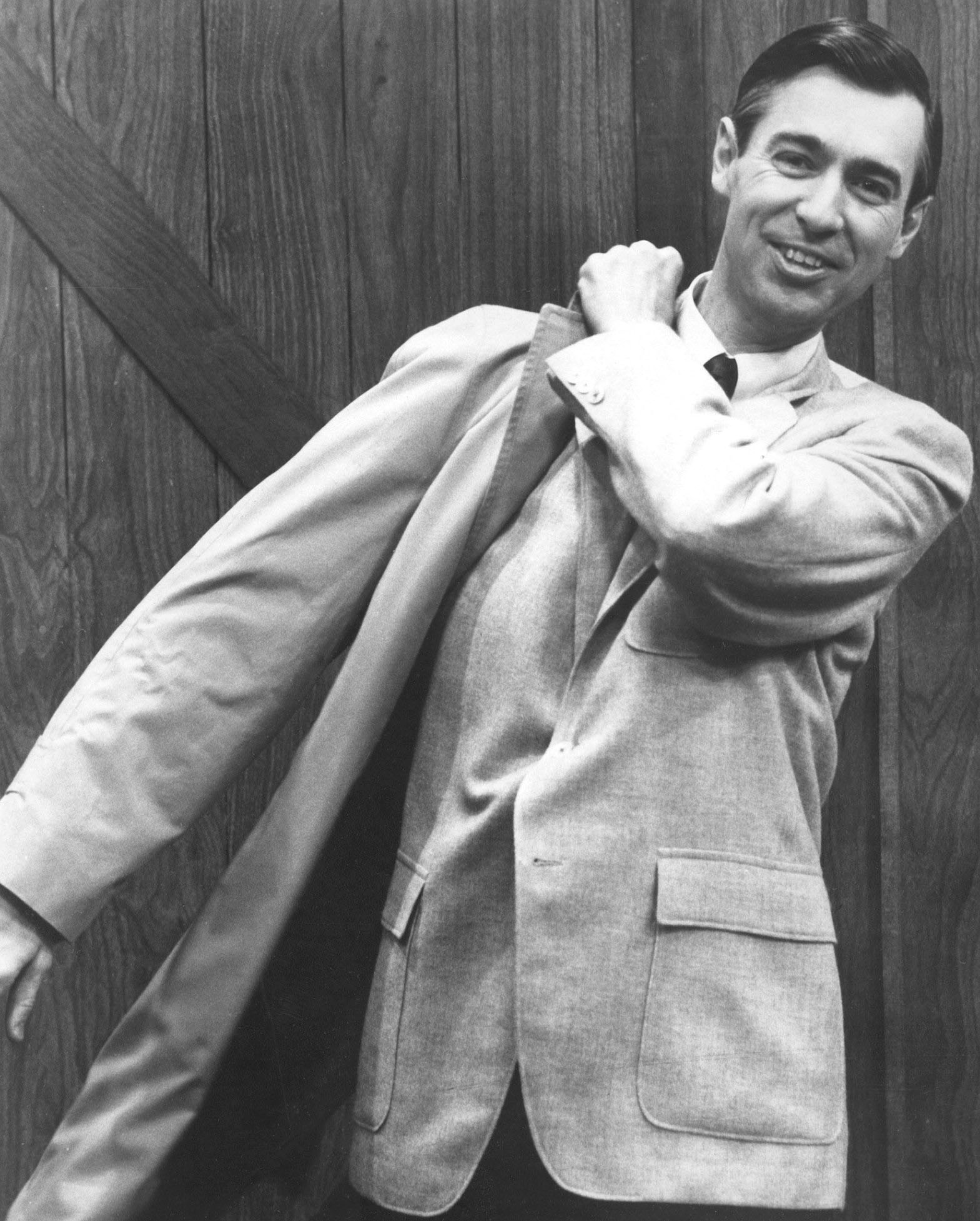 A grayscale half-body photograph of Fred Rogers putting his coat on to bid his audience goodbye at the end of an episode of the television show Mister Rogers' Neighborhood.