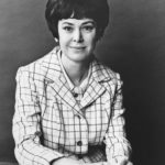 Grayscale half-body photograph of Joan Ganz Cooney from 1968. She is facing the viewer head-on, wearing a button-down blazer with tattersall checks, with hear hands slightly crossed on the table in front of her.
