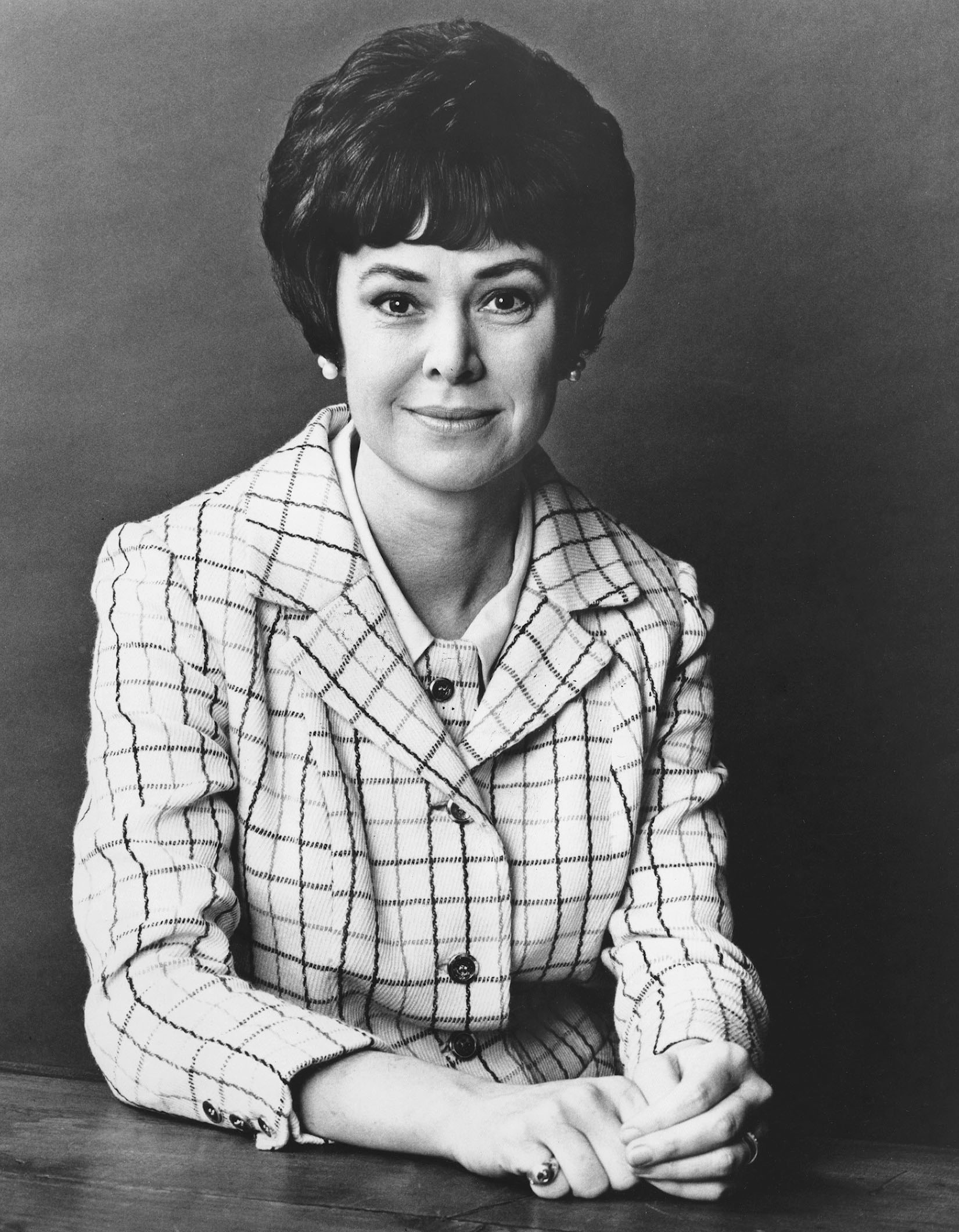 Grayscale half-body photograph of Joan Ganz Cooney from 1968. She is facing the viewer head-on, wearing a button-down blazer with tattersall checks, with hear hands slightly crossed on the table in front of her.