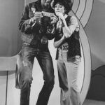A grayscale full-body photograph of Morgan Freeman and Rita Moreno standing closely together to examine a dollar bill in an episode of the television show The Electric Company.