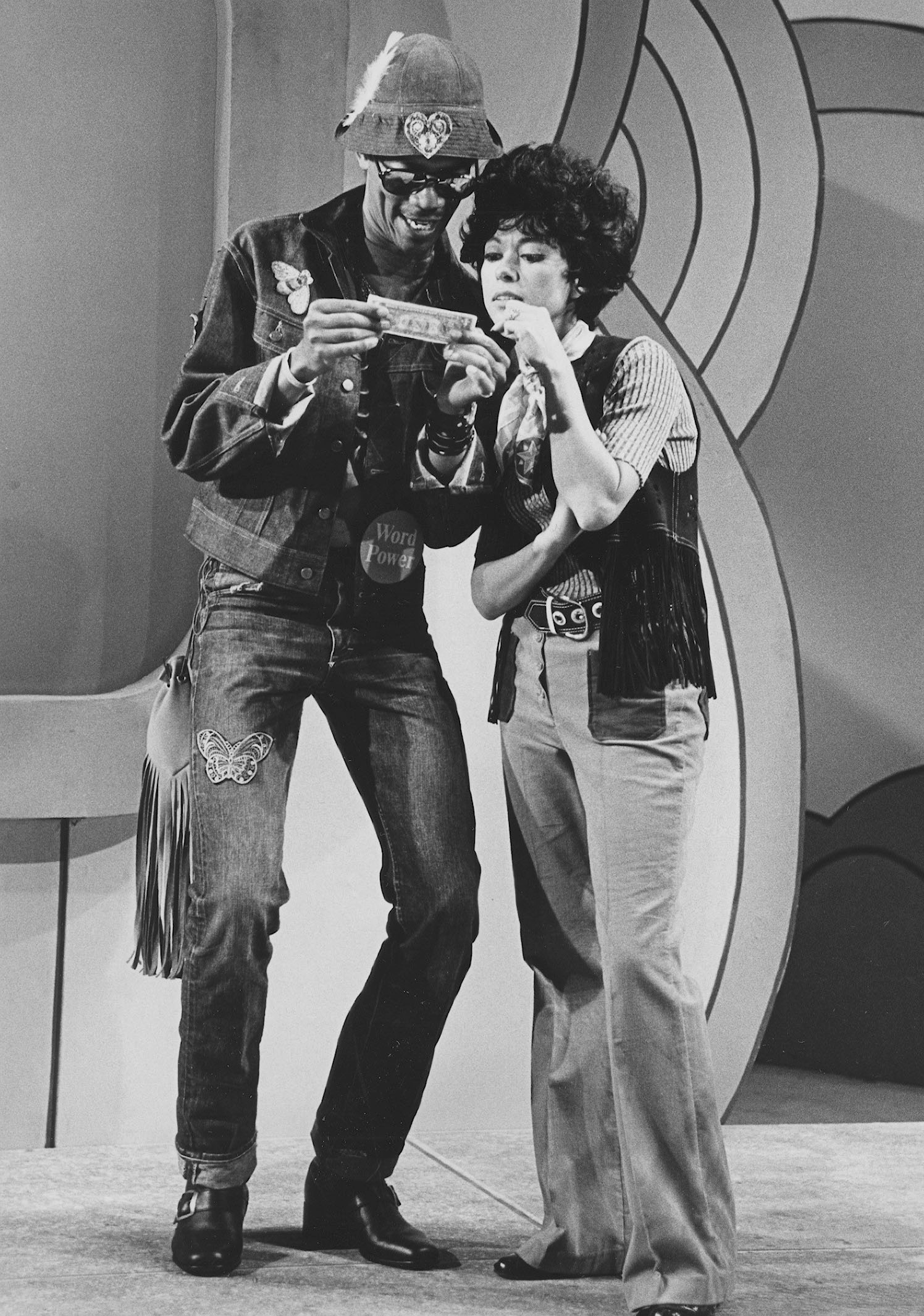 A grayscale full-body photograph of Morgan Freeman and Rita Moreno standing closely together to examine a dollar bill in an episode of the television show The Electric Company.