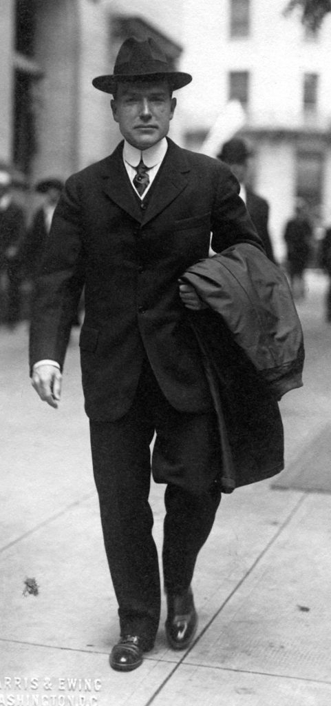A portrait of John D. Rockefeller, Jr., dressed in a dark three-piece suit and hat, walking on a city sidewalk.