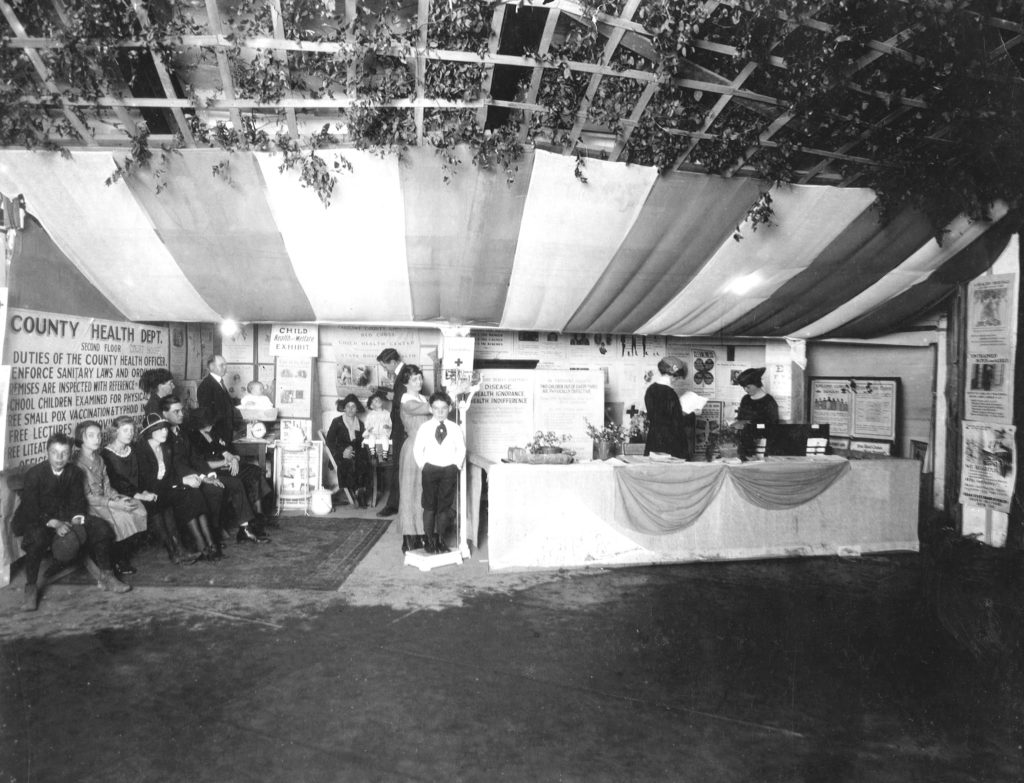 People gathered in a booth at a public health demonstration at a county fair in Texas in the 1900s. They are looking at an exhibit about hookworm disease.