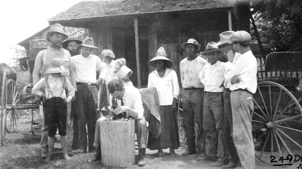 A group of people at a public health dispensary for the treatment of hookworm disease, gathered around a man who is looking into a microscope.