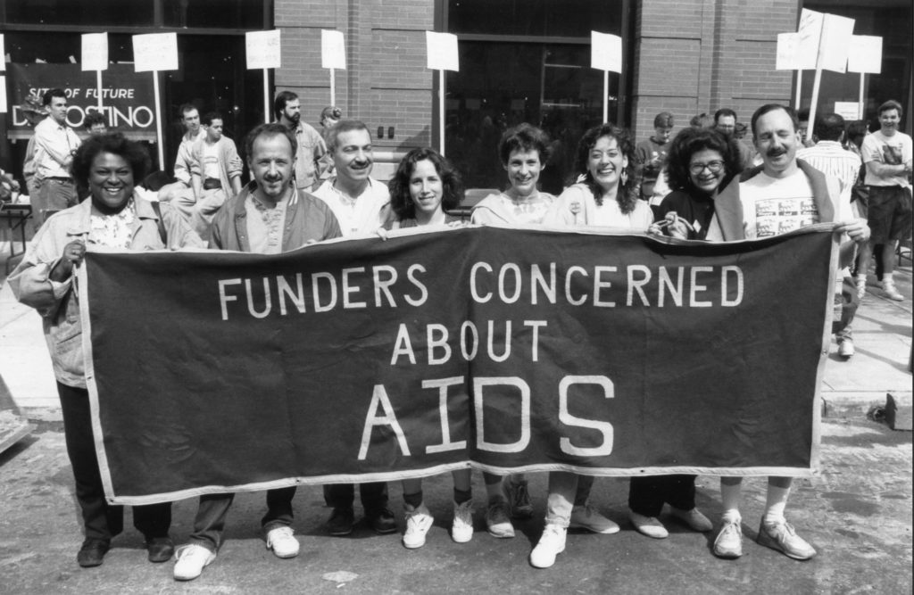Eight people of different races and genders hold up a banner that says "Funders Concerned About AIDS," with people marching and holding signs in the background.