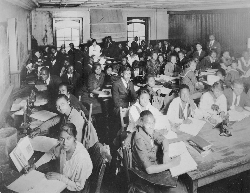 A classroom photo of African American college students, both men and women, at Shaw University in North Carolina. The students sit at tables and are looking at books and taking notes in notebooks. Work in black education spanned elementary schools to colleges and universities.