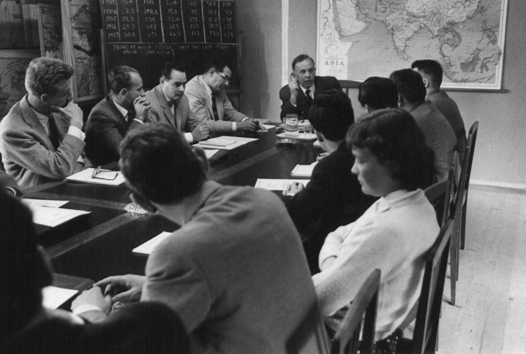 Several men and one woman all in formal dress sit around a table, with a map of Asia and a chalkboard behind them.