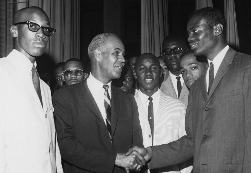 NAACP leader Roy Wilkins shakes hands with another Black man, surrounded by several other Black men. All are wearing suits and ties.
