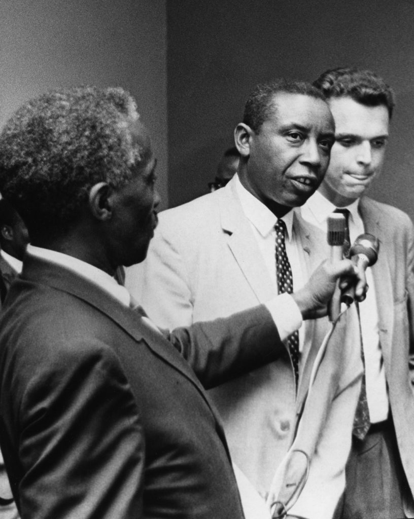 Congress of Racial Equality president Floyd McKissick, a Black man, speaks into a microphone held by another Black man. A white man stands to McKissick's left.
