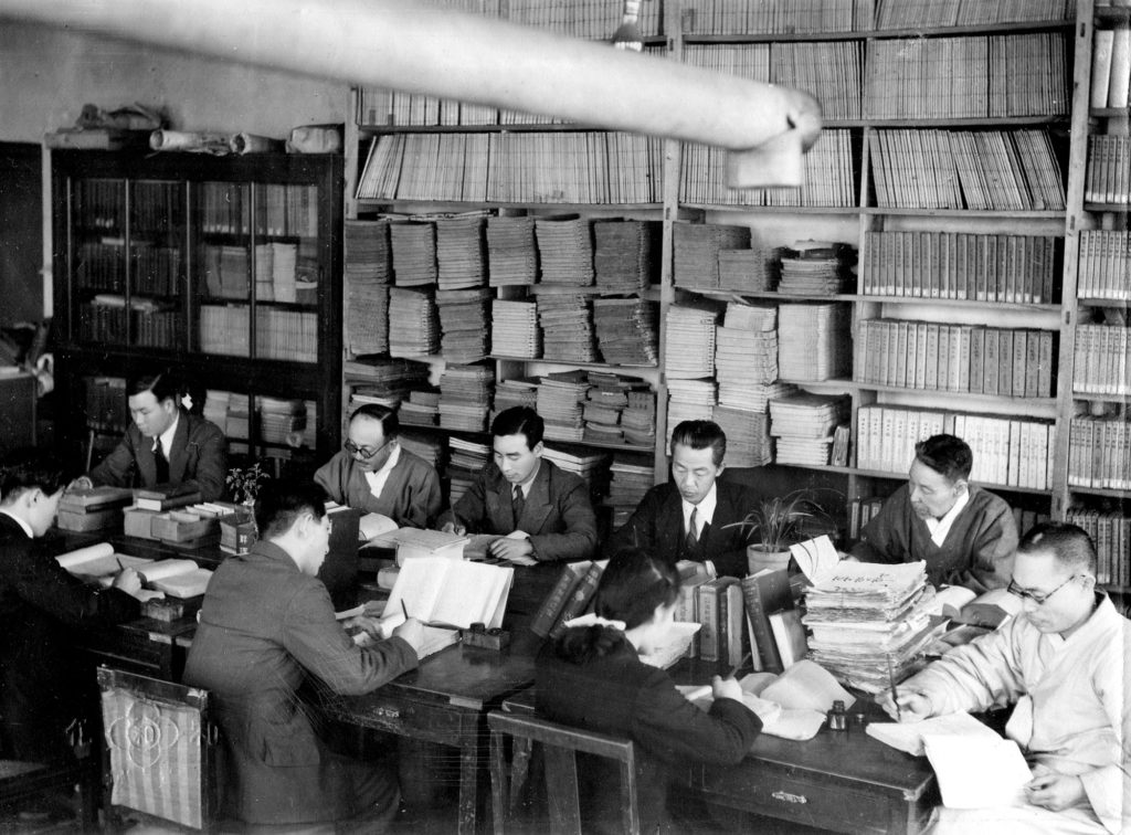 Seven Korean men and one woman work at a large table covered in papers and books. They are working on the Korean (Hangul) Dictionary.