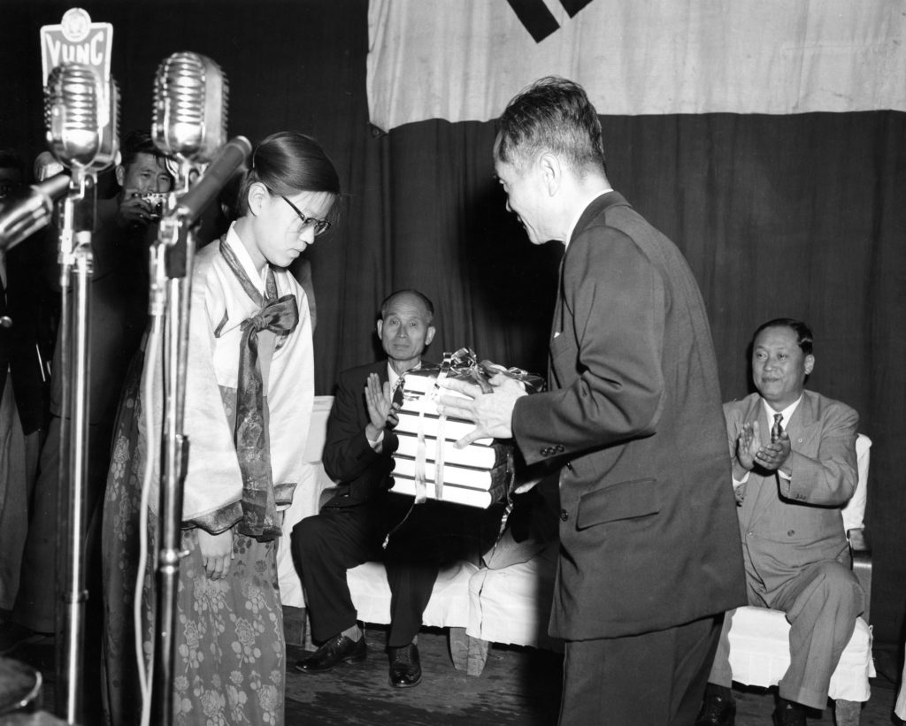 A Korean woman in traditional silk robes stands at left while a man at right holds the six-volume Korean (Hangul) dictionary, wrapped in ribbons.