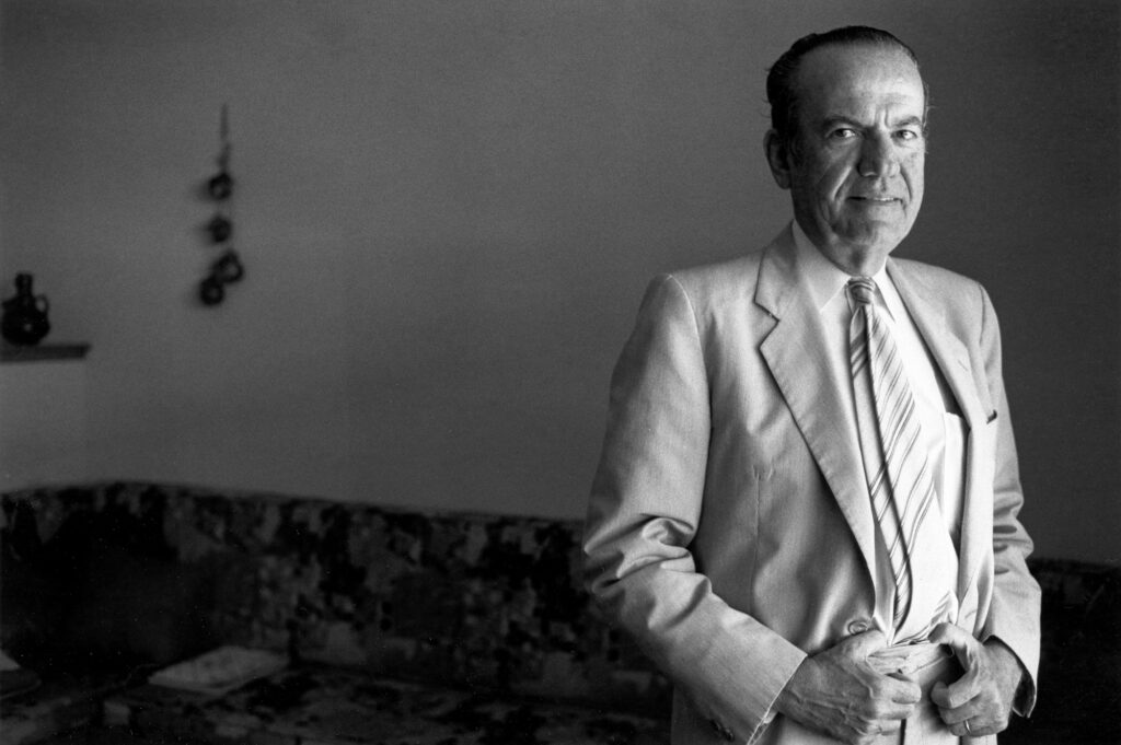 Black and white portrait of Dr. Jose Barzelatto dressed in a suit and tie and standing in a sitting room.