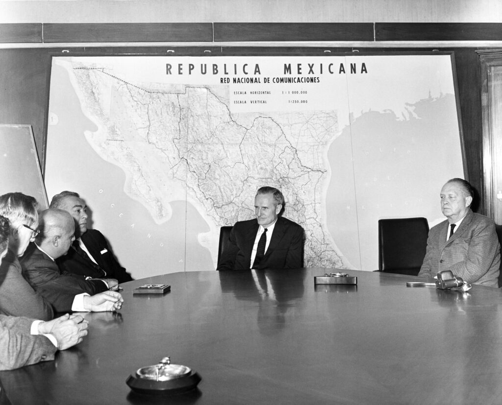 A grayscale photograph of E.J. Wellhausen, Jose Rodriguez Vallejo, Ricardo Acosta, John D. Rockefeller 3rd and J. George Harrar seated around a table with a shiny surface. A large scale map of the national communications network of Mexico is displayed behind the seated men.