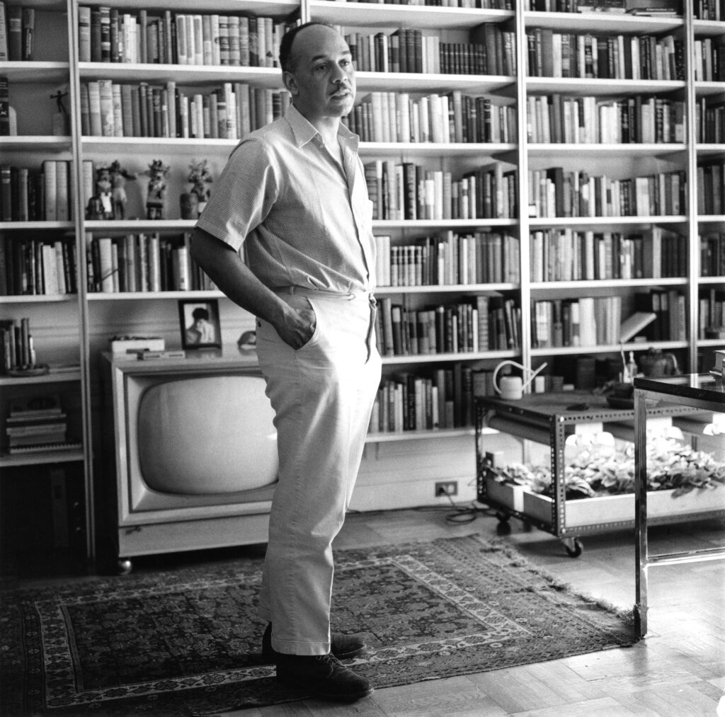 A full-body shot of Ralph Ellison standing in a room, with a television box and bookcase in view.